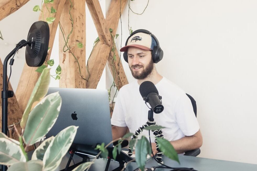 Man podcasting with laptop and microphone, surrounded by indoor plants and wooden decor.