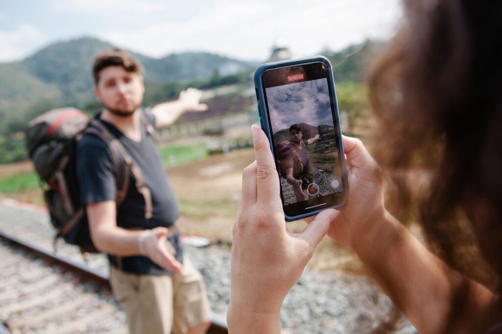 Crop unrecognizable female taking video on smartphone of hiker in casual clothes with backpack standing on railroad and pointing away in countryside in sunny day