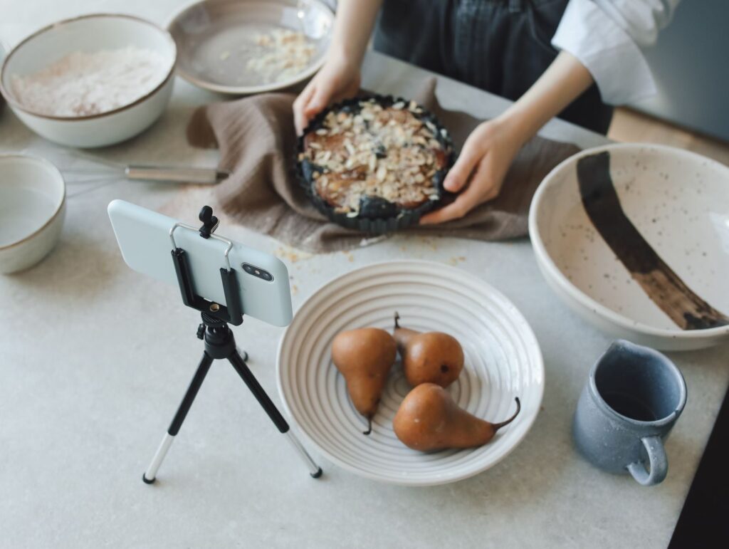 Woman baking in kitchen while recording with smartphone, showing delicious pears and a cake.