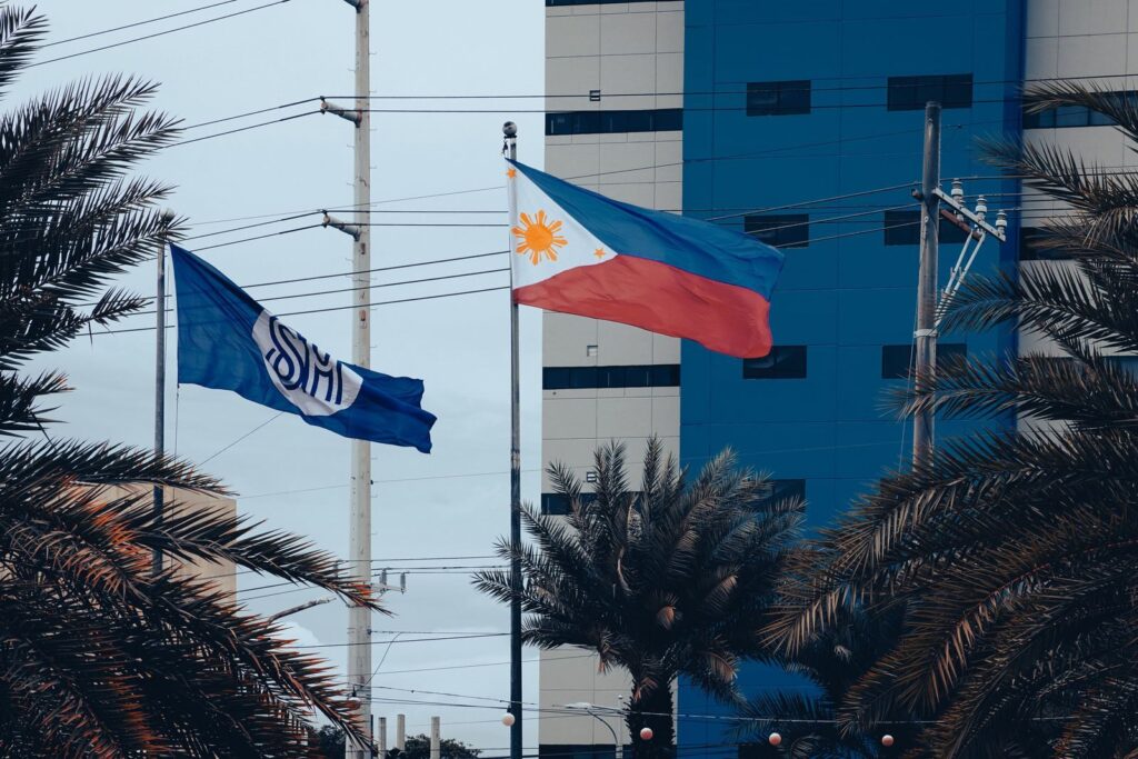 Philippine and SM flags flutter outside a contemporary building in Manila with palm trees in foreground.