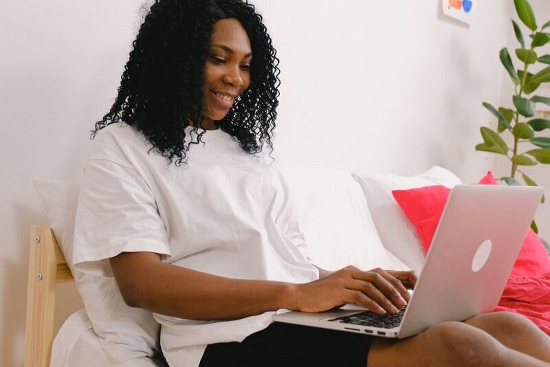 Positive African American female typing on netbook while sitting on soft bed near wall with colorful decorations in bedroom with green plant